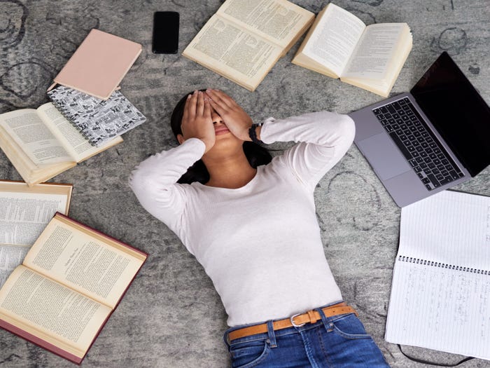 A girl lying on the floor surrounded by schoolwork with her hands over her eyes.