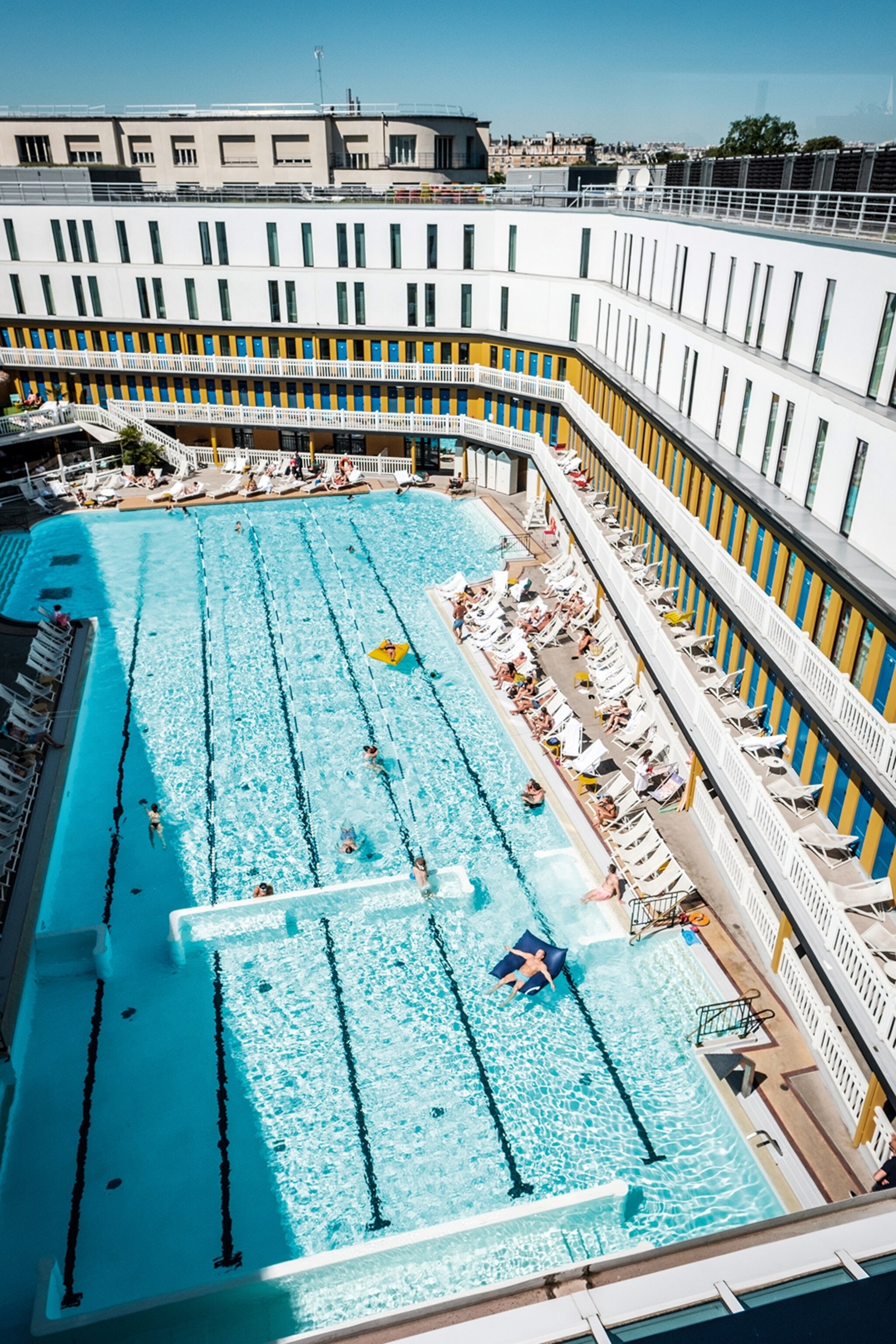 A swimming pools in Paris, some people in in the water, whilst some dangle their legs in from the side.