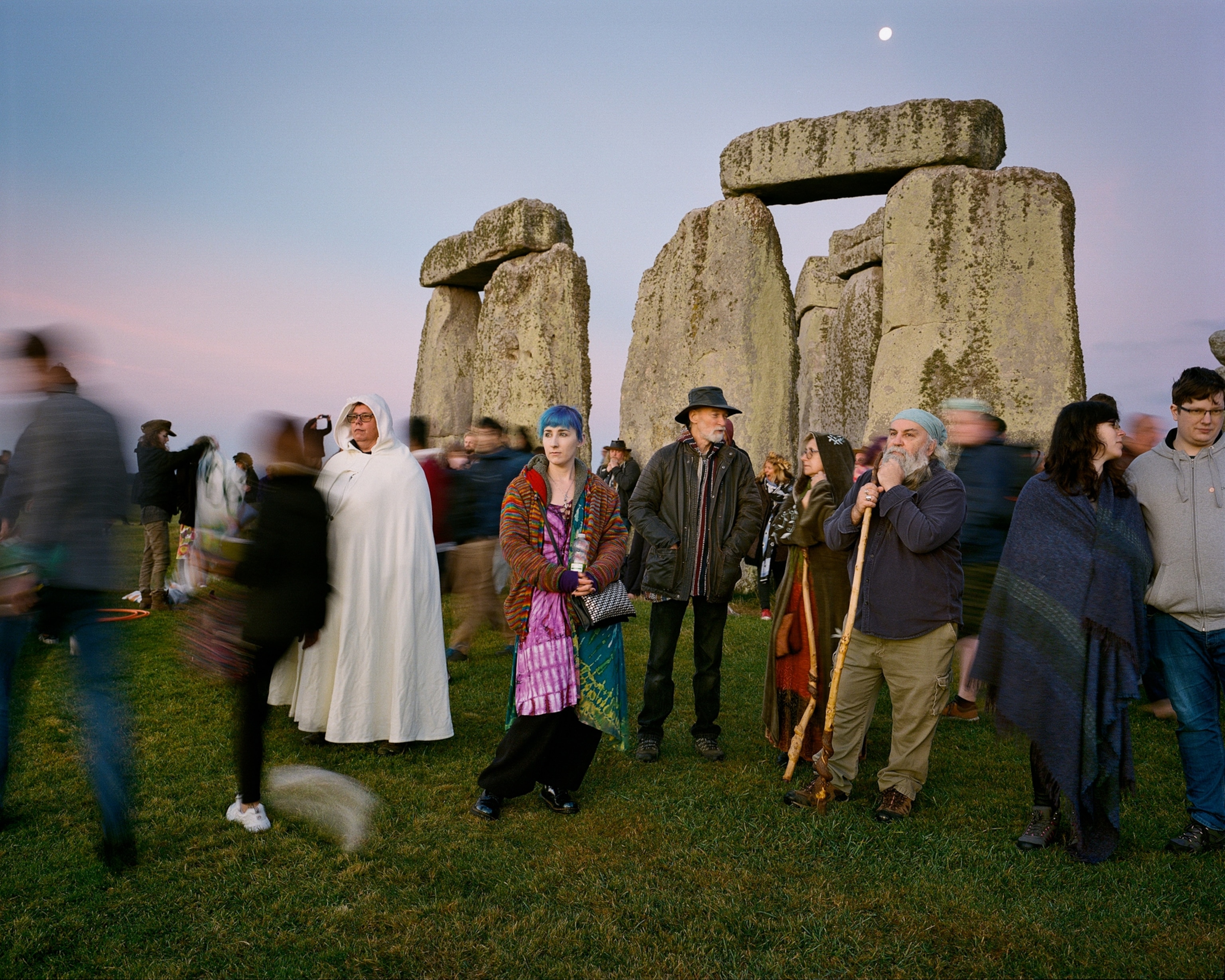 An eclectic crowd of people gathered by the ruins of Stonehenge with a full moon visible in the sunset sky.