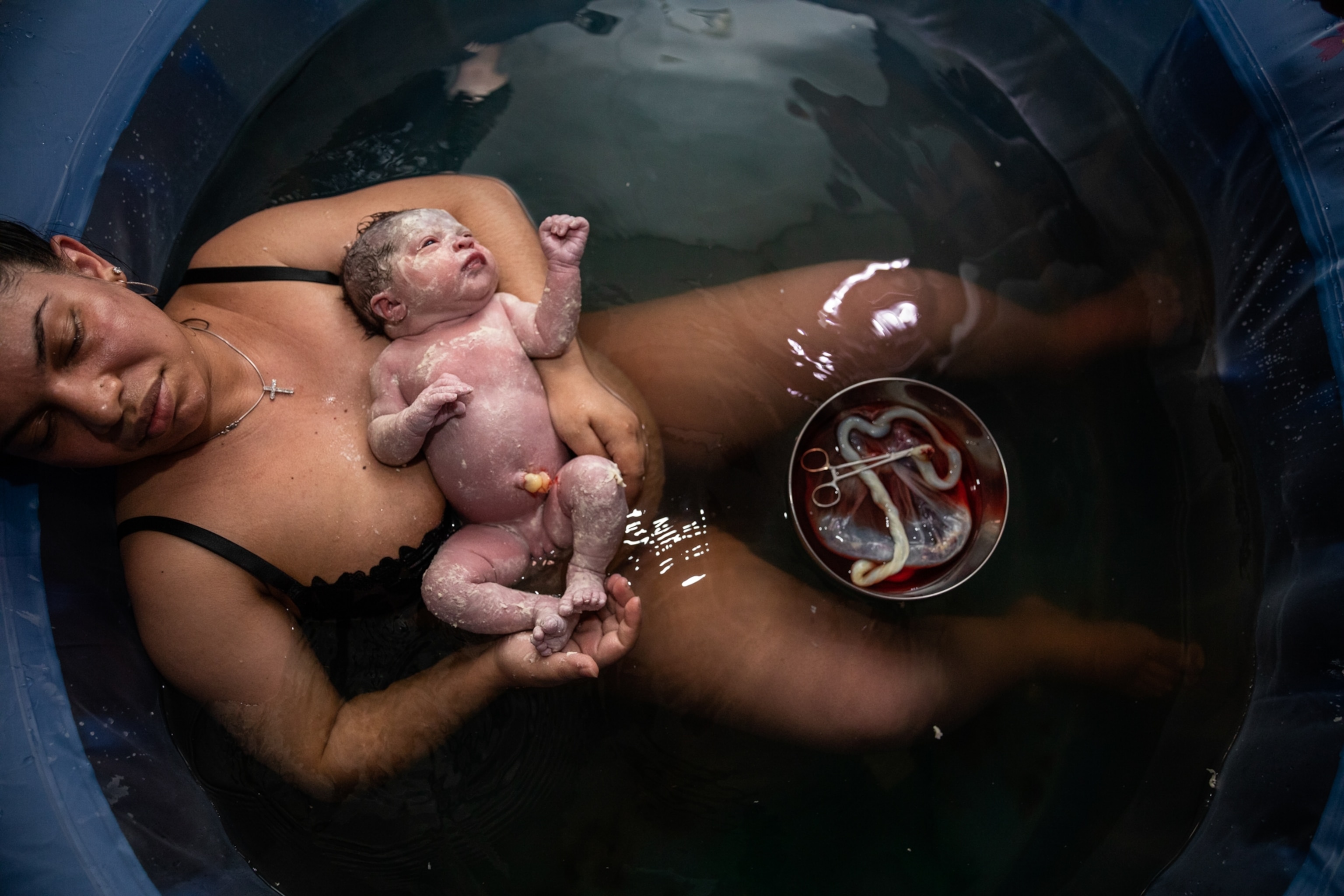 A woman lies back in a small birthing pool, her newborn daughter cradled in her arms.