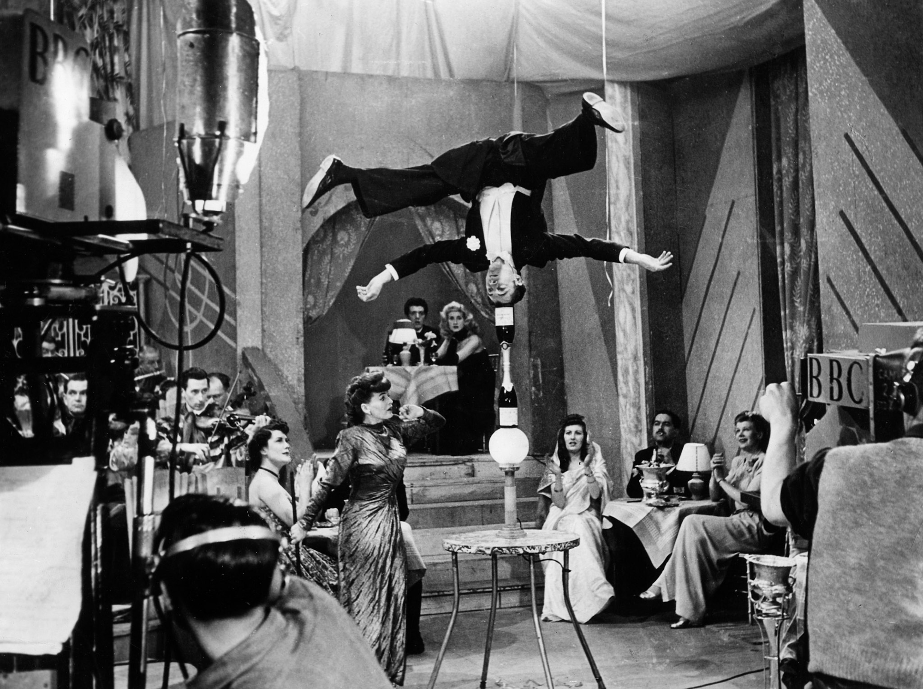 Man surounded by audience and camera black and white image. Man is doing headstand balancing act.