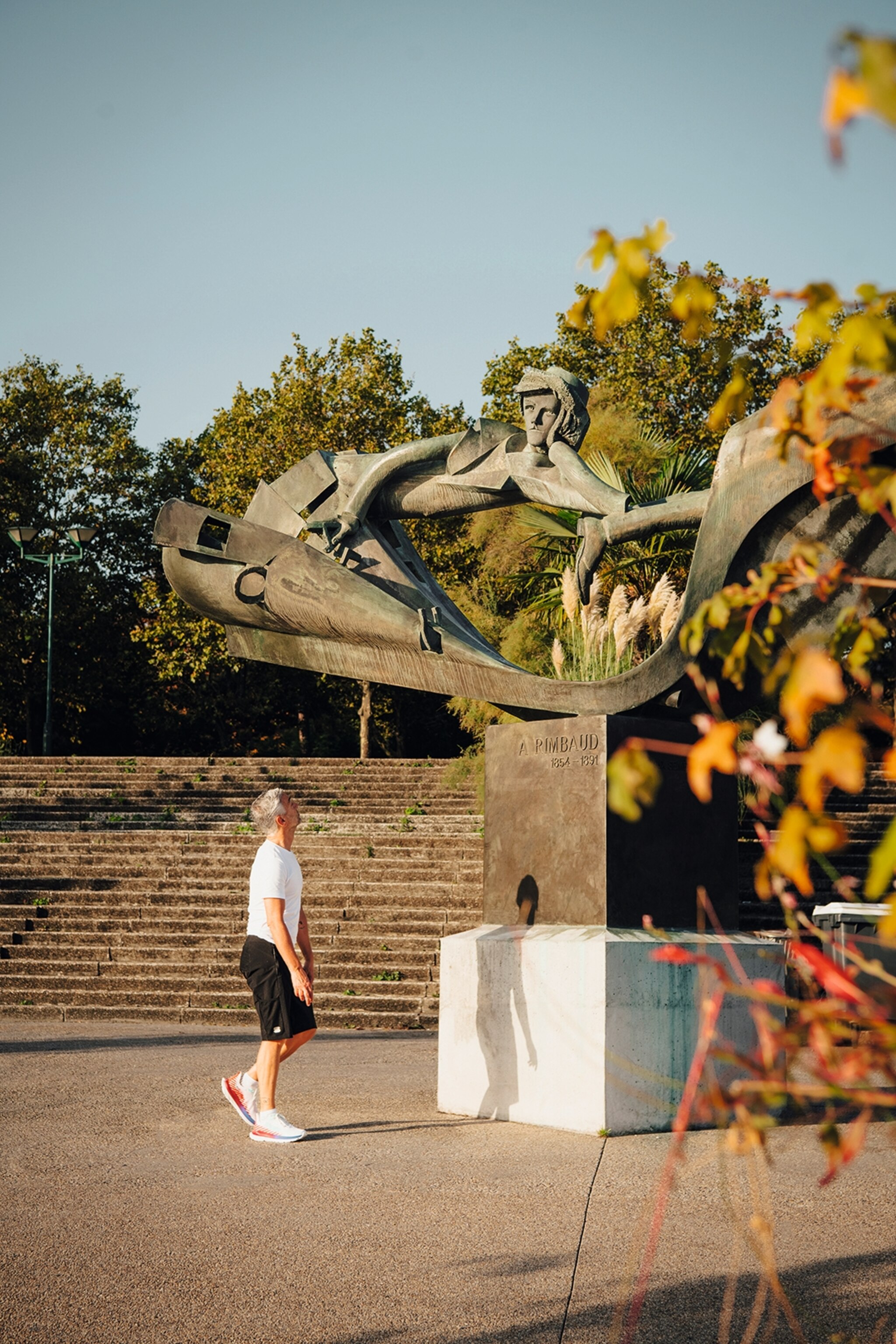 A runner wearing a white tee, blakc shorts and sneakers looking up at an outdoor stone sculpture.