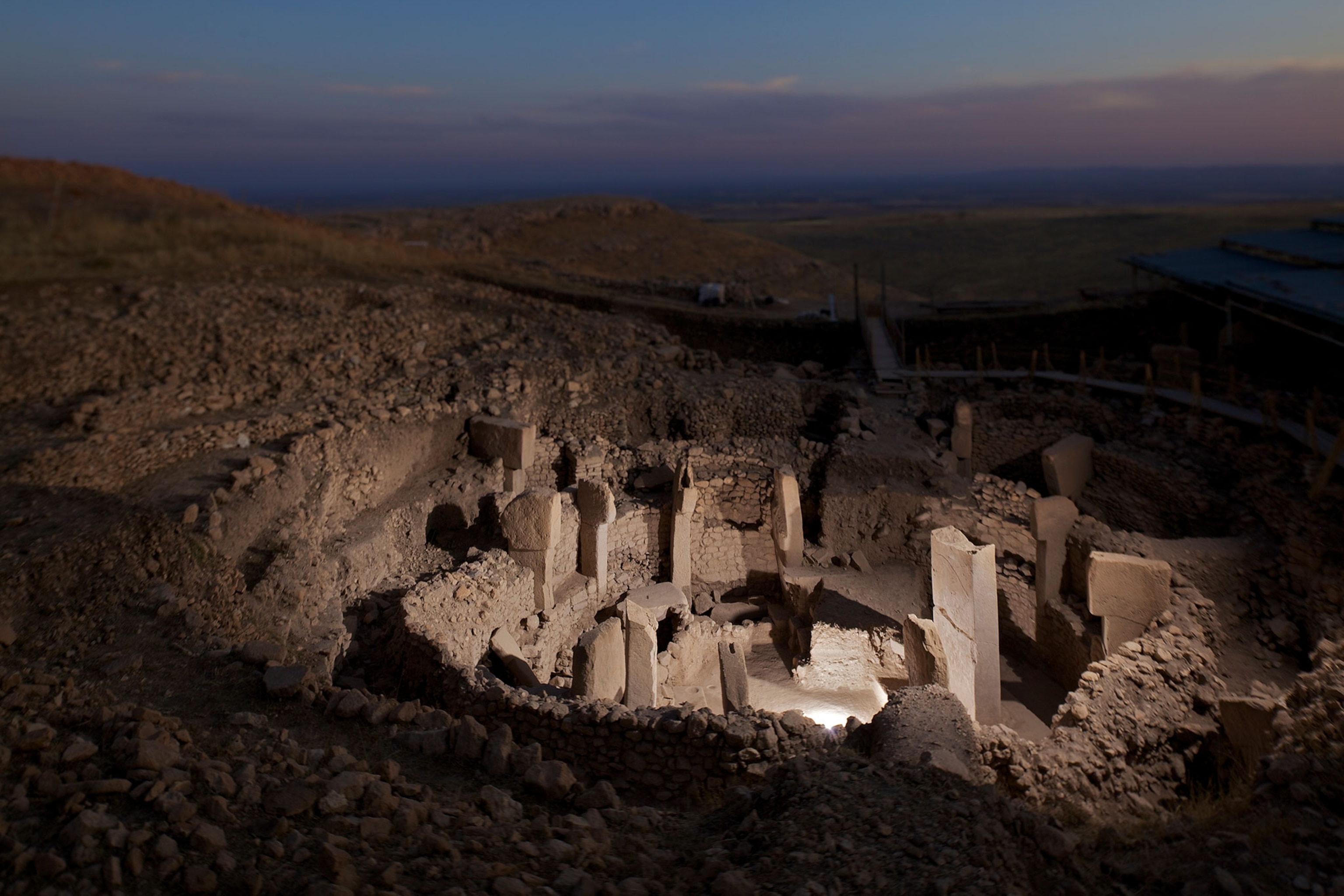 Looking down at Gobekli Temple, there are old stone pillars standing in a circular shape. The blue and pink sky is seen behind the temple as the sun sets. A bright light coming from within the temple lights up the site at dawn.