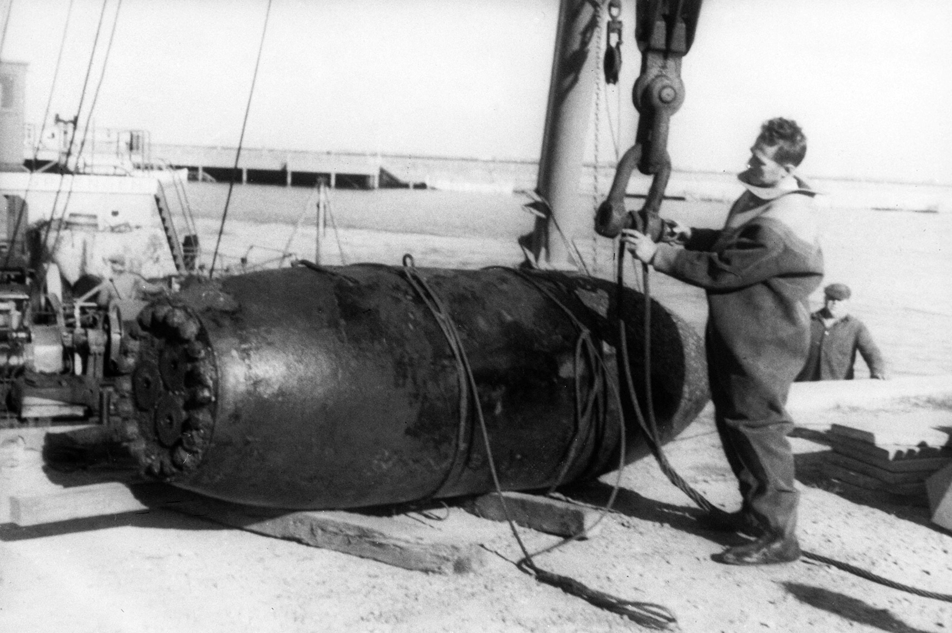 A man in a workman's jumpsuit, connects a large, thick wire to a crain. The wire is wrapped around a thick, black, bomb, about three times the size of the man.