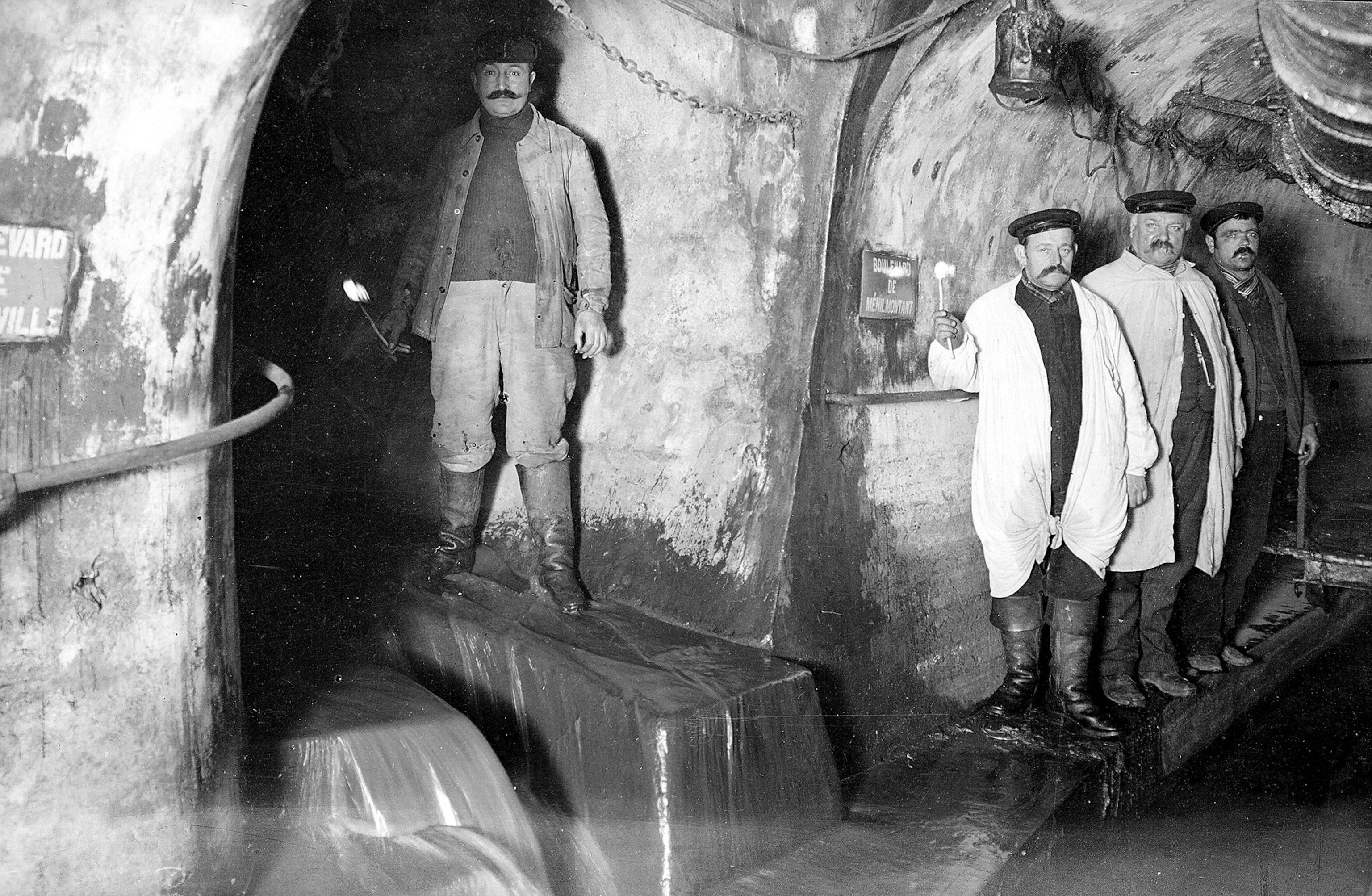 a black and white from the 1890s of men standing in an underground sewer with water flowing