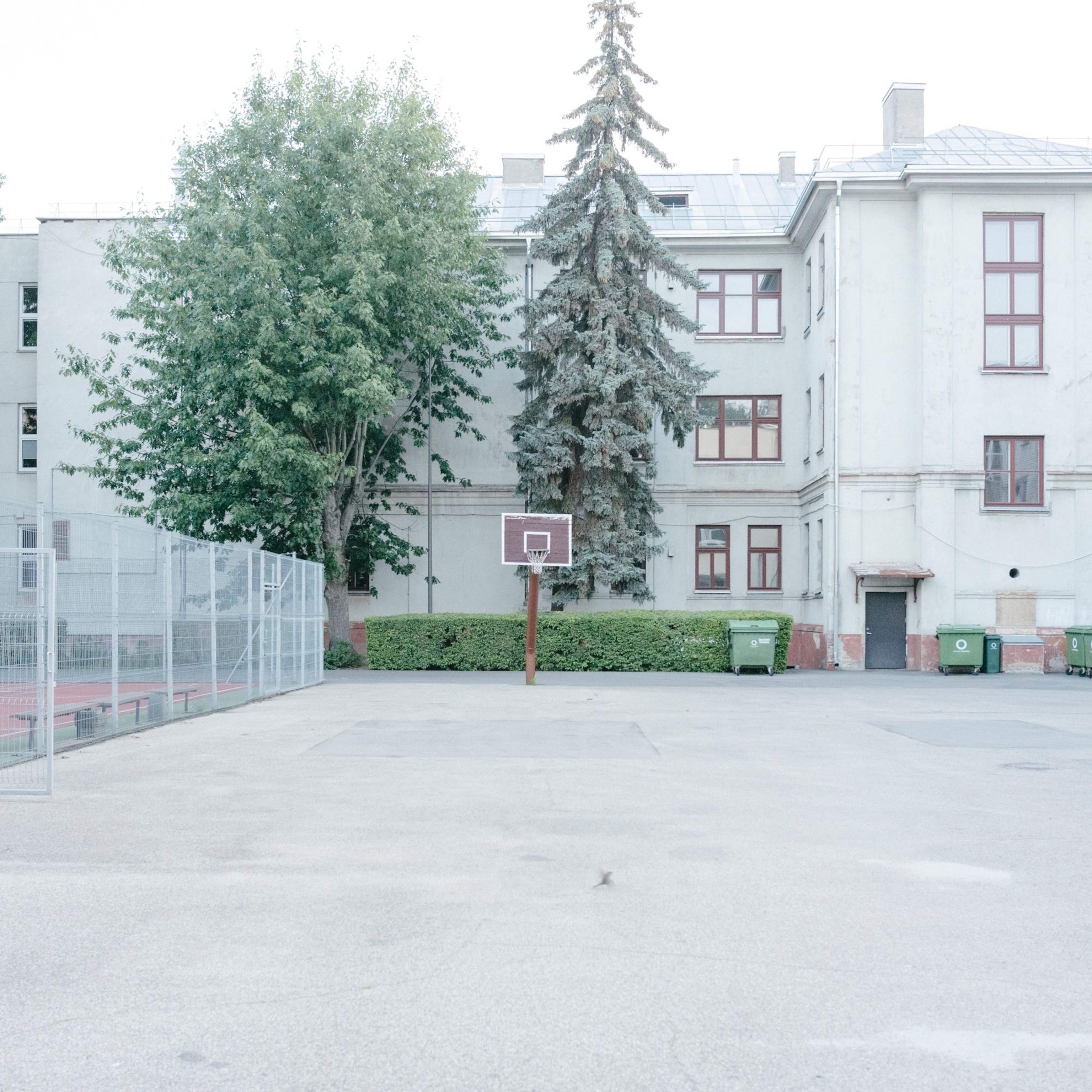 An image of a concrete basketball court next to a building with modern recycling bins