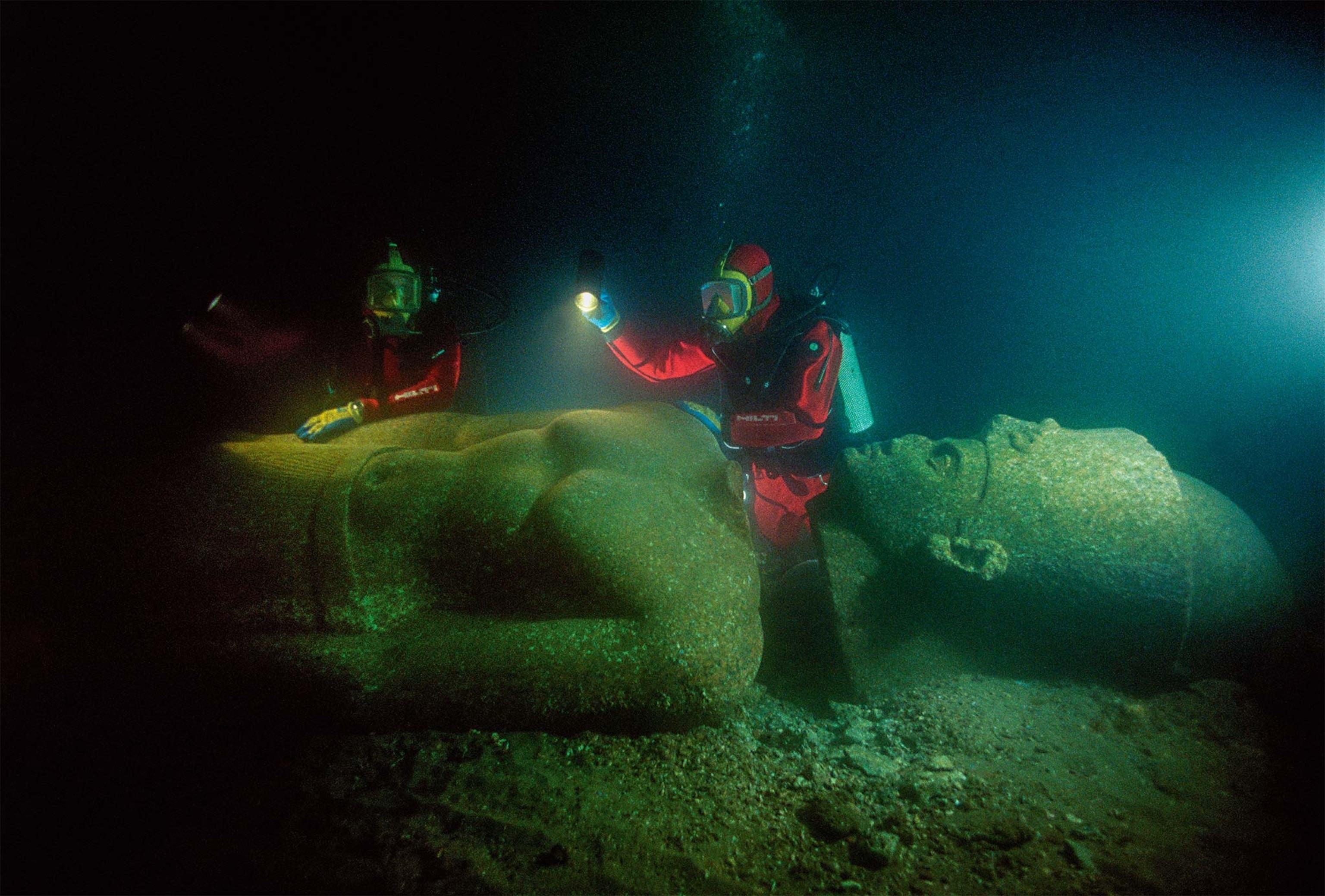 Divers in red suits look at a sunken god statue in the ocean