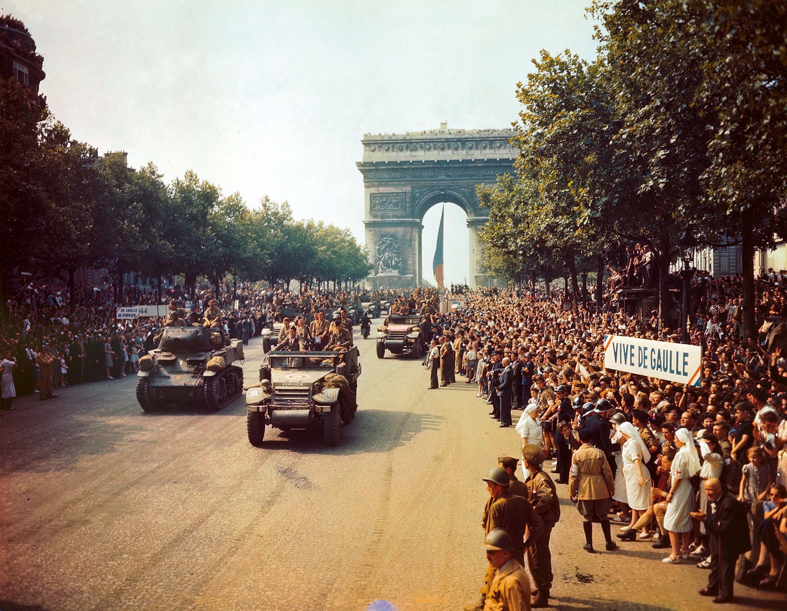 French military vehicles parade in front of the Arc de Triomphe among crowds celebrating on the street