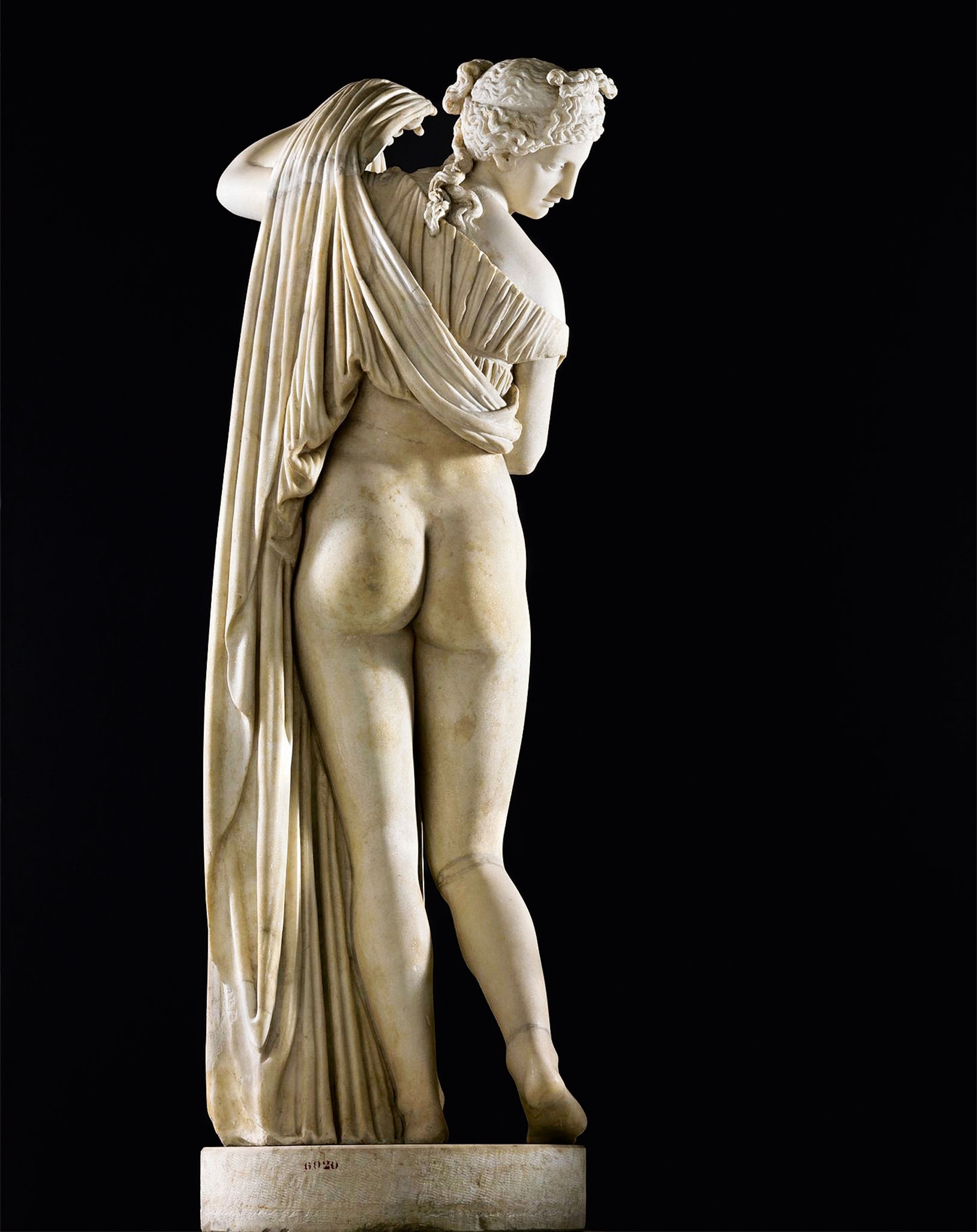 A stone statue of a half-naked woman from the back, with a draped fabric covering her top half