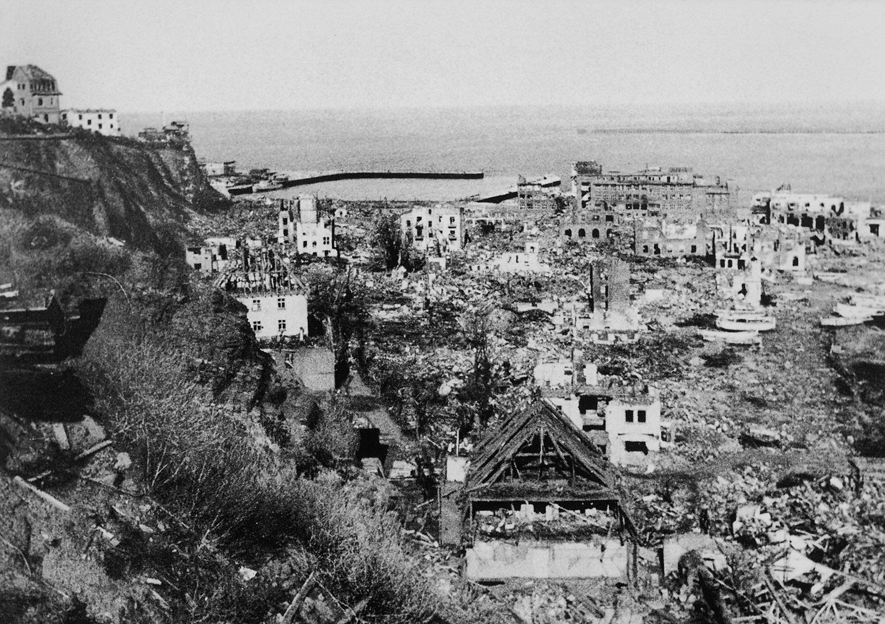 A picture of a town, decimated by a bomb. Buildings are in shambles and debris is everywhere. The town borders the sea at the top of the photo and a cliff to the left.