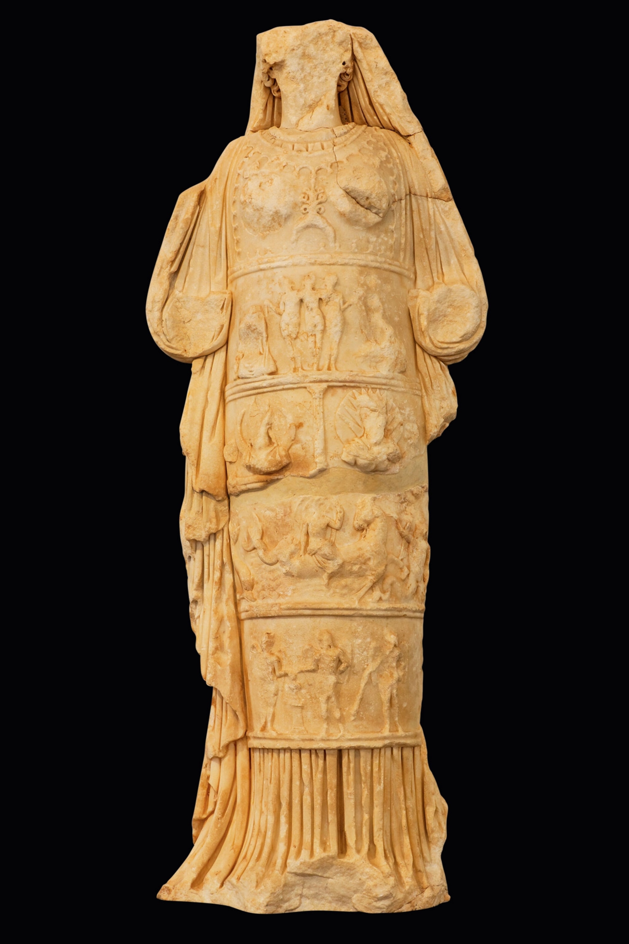 A stone statue, missing its face, of a woman in a dress with layers of carvings