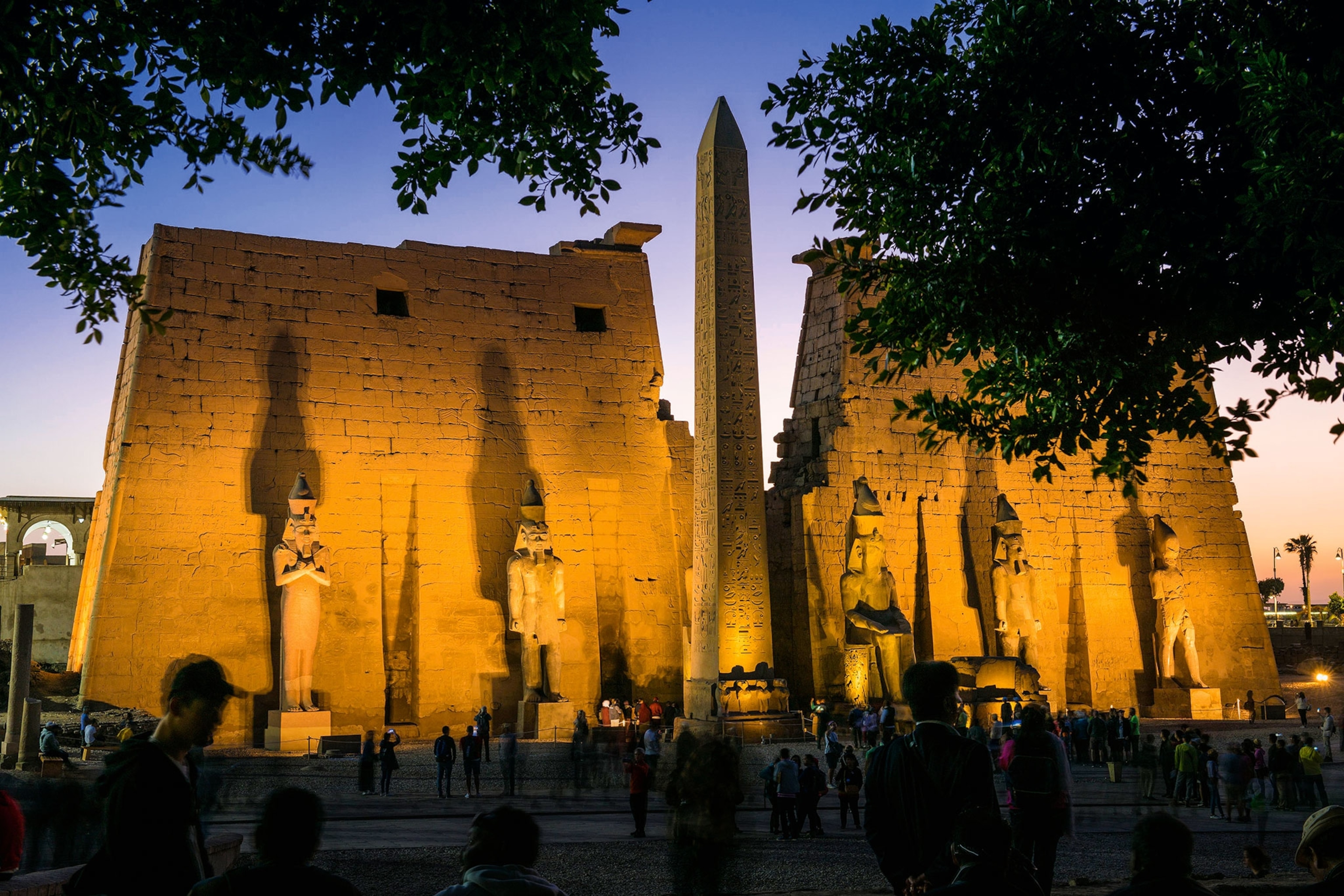 A wall with statues in front of it is illuminated, with a break in the middle revealing an obelisk