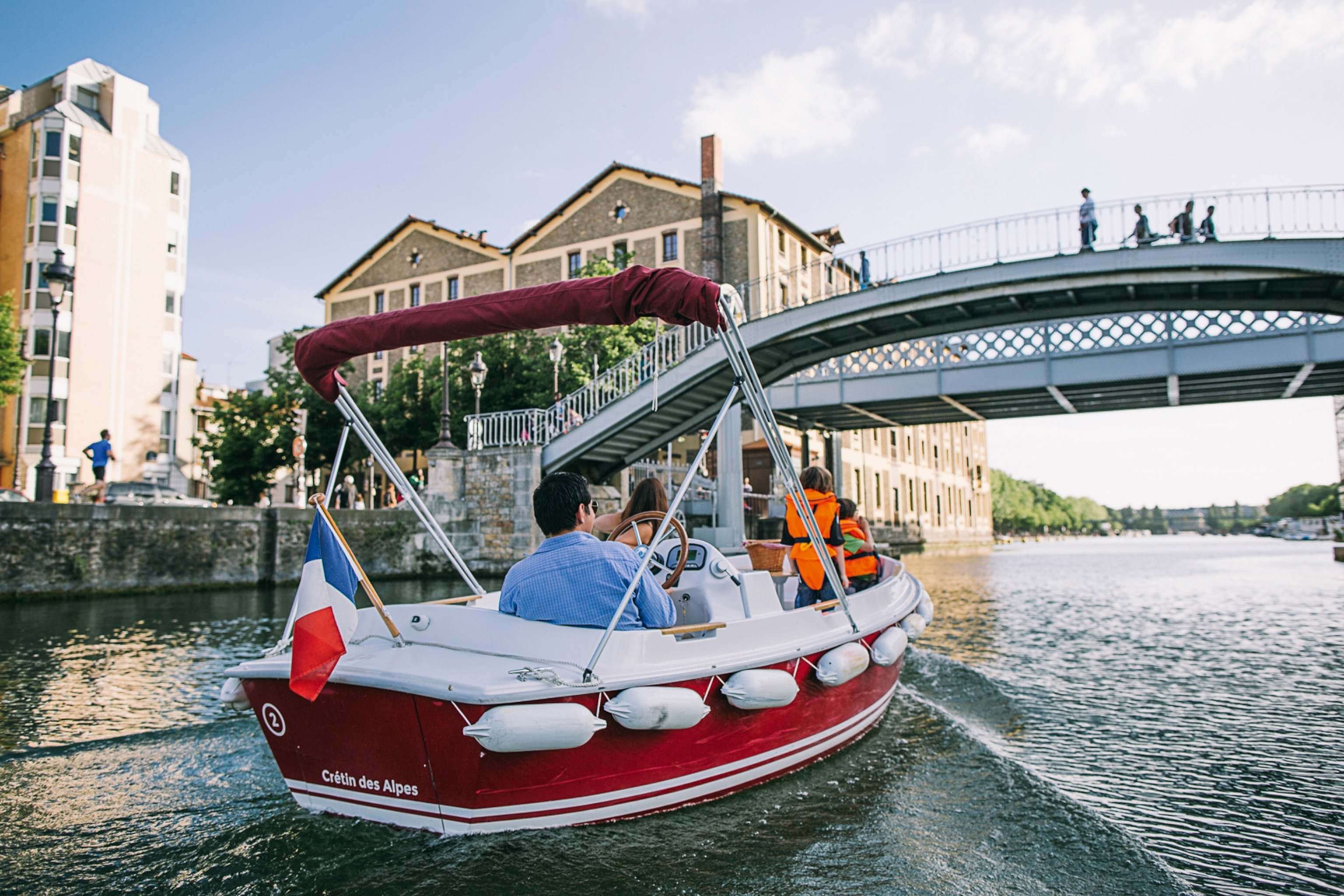 People taking a boat trip down a lake. It is a sunny day and the water is calm. People on the bridge above are stopping to look out at the water.