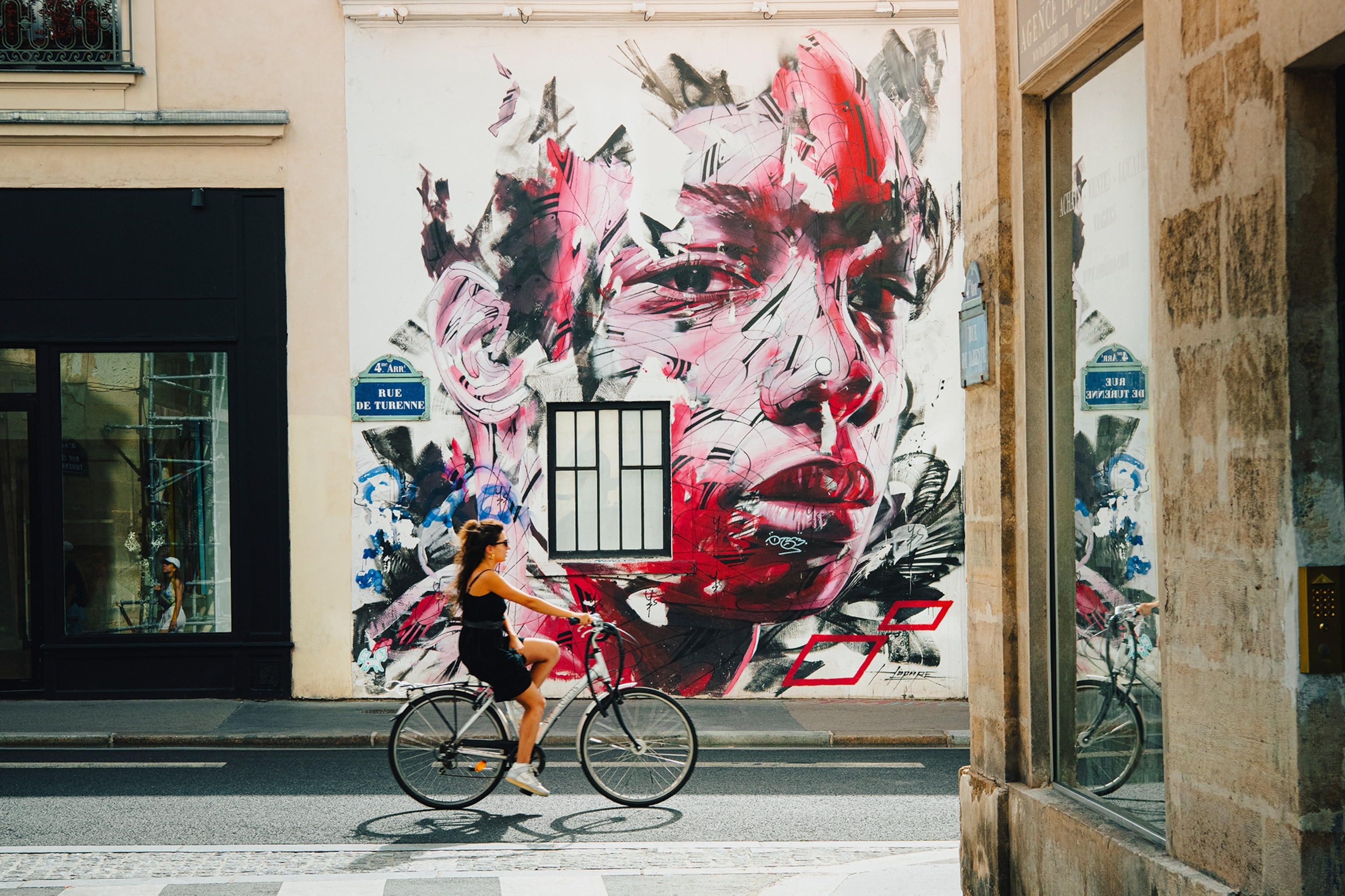 A woman in a black outfit is riding a bike in Paris, the wall beside her has an abstract graffiti design