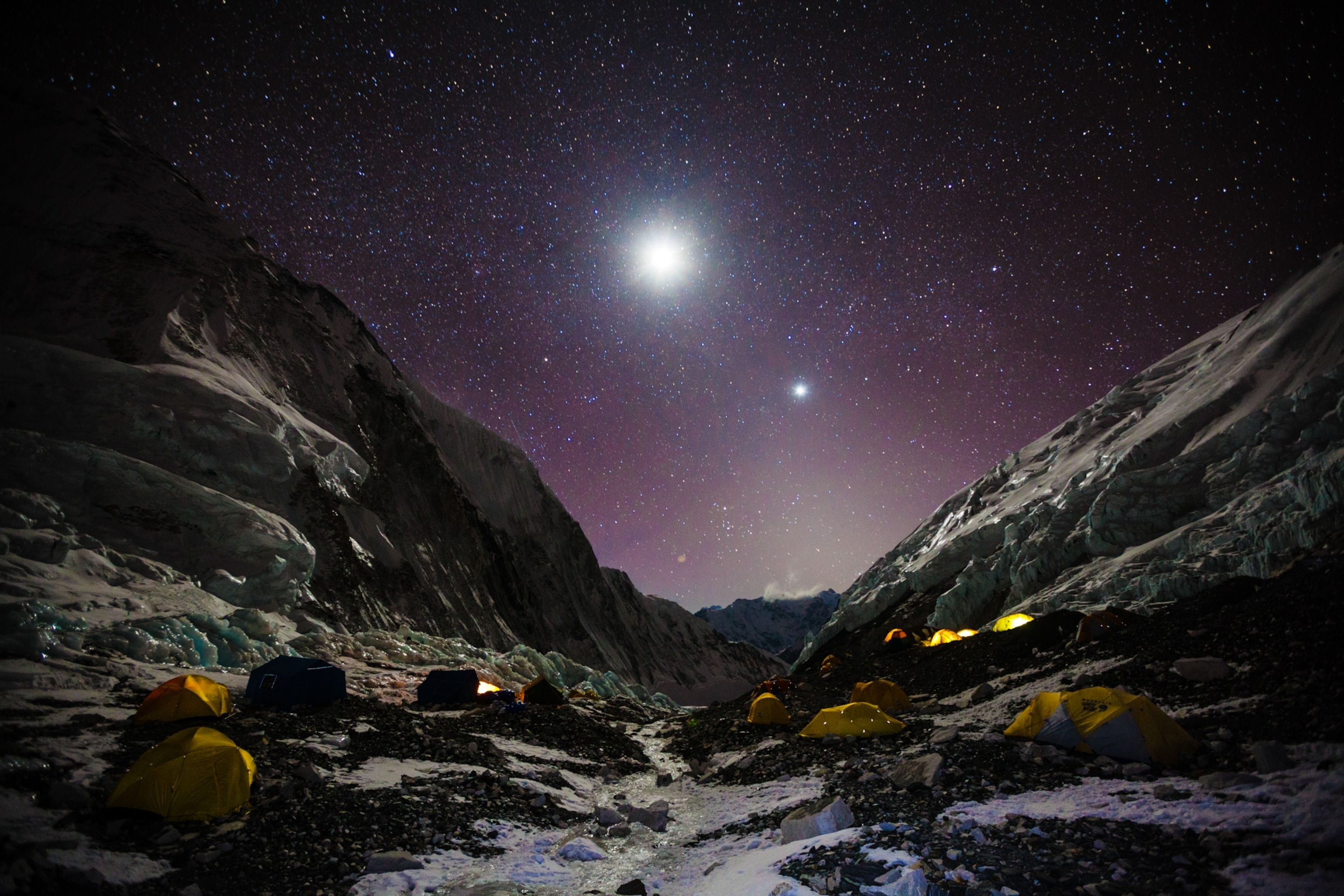 A star-filled sky and bright moon shine over dozens of tents set up in at a rocky and icy campsite on Everest.