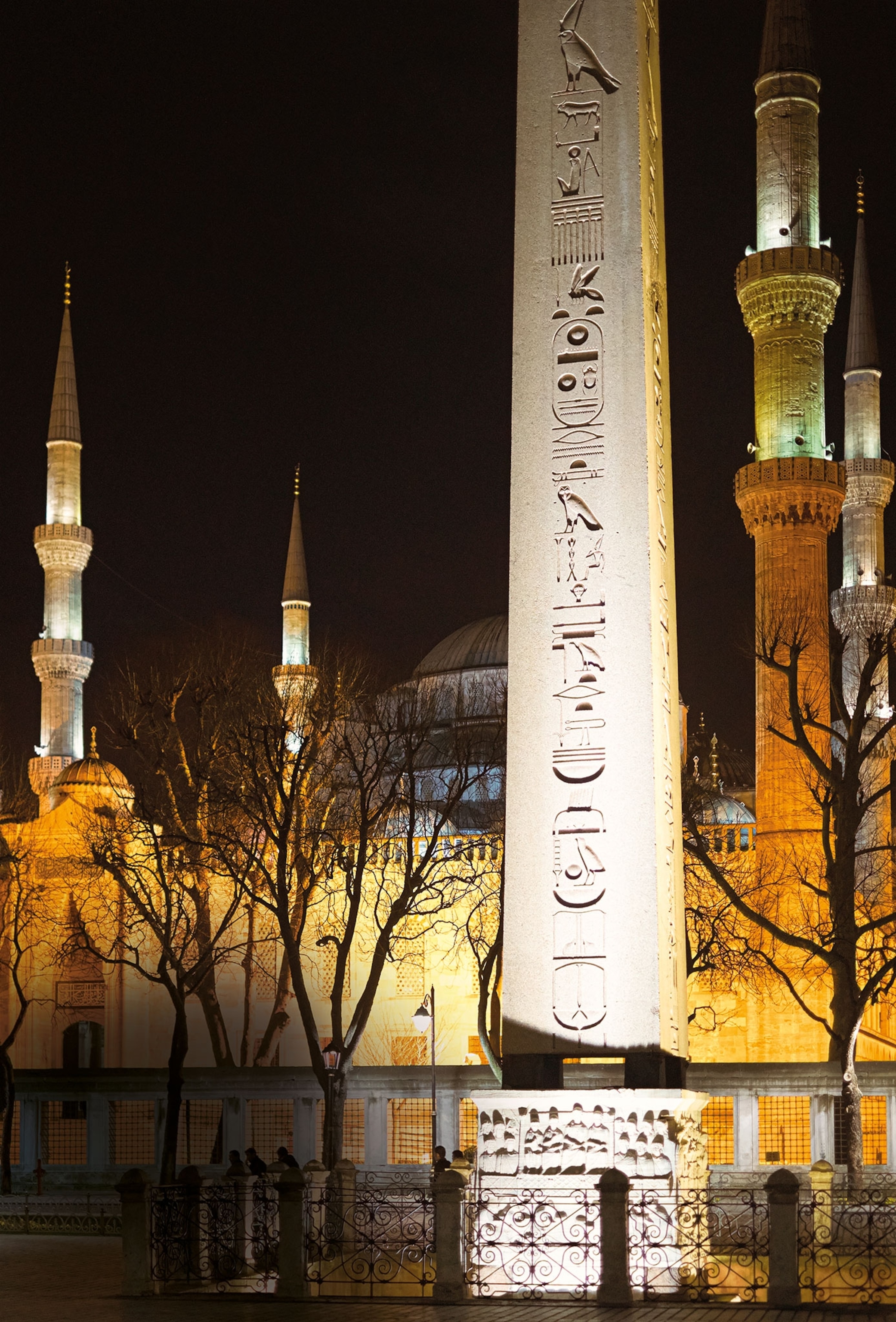 Minarets of a mosque appear behind an obelisk at night