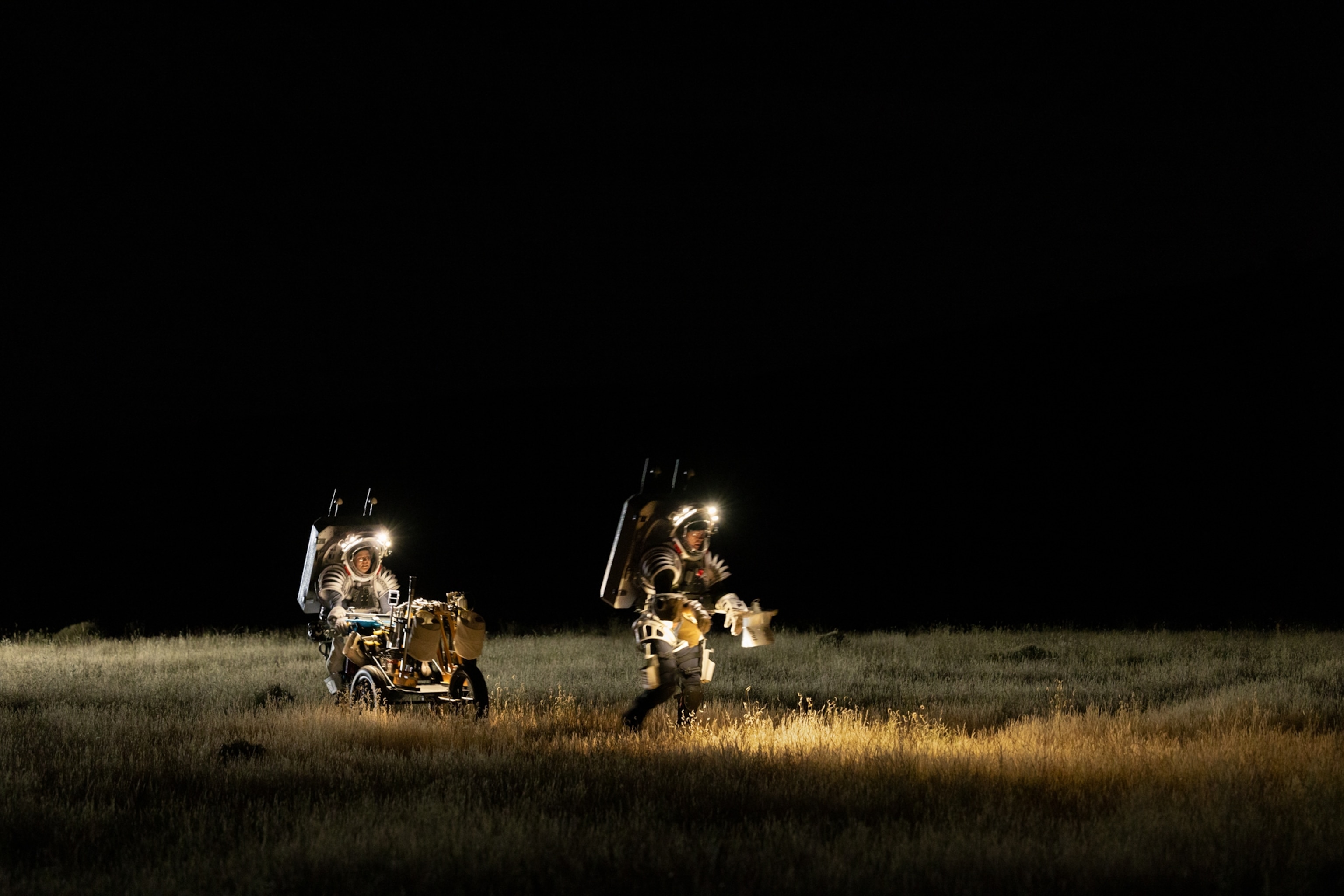 Two people in full space suits with headlights working on the field at night. One with the heavy loaded stroller.