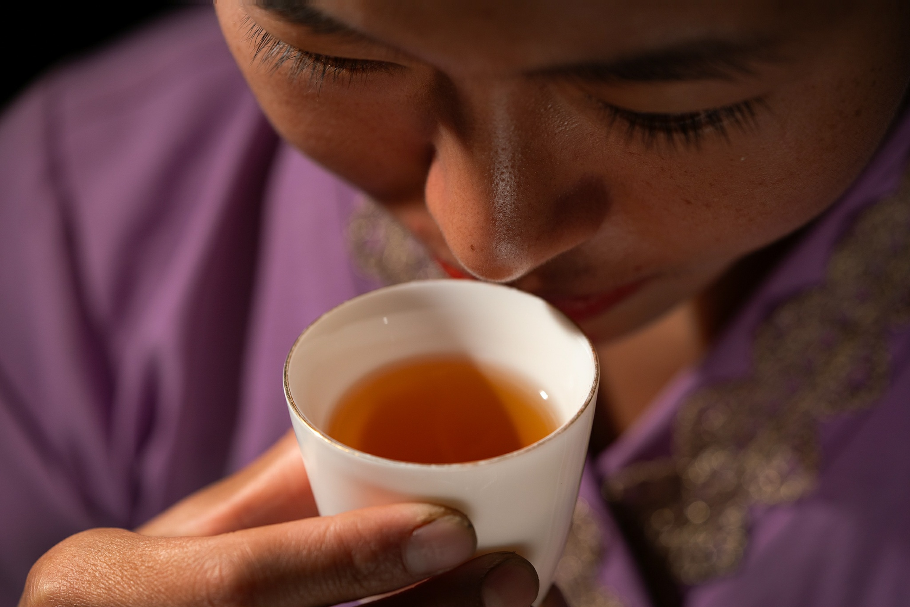 A close up view of a woman lifting a small white cup of amber colored tea up to her nose.