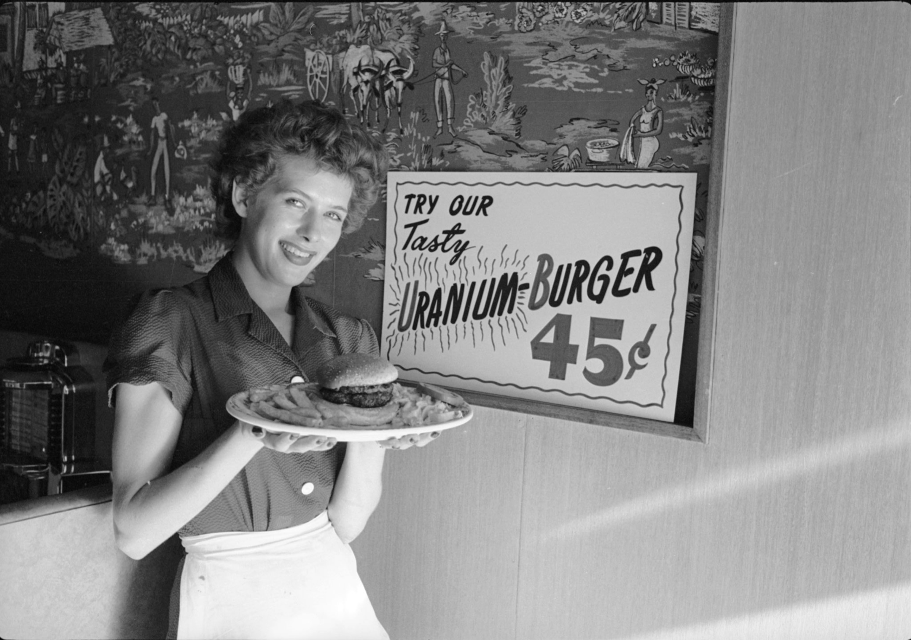 A black and white photo of a waitress poses with a 'Uranium-Burger' at a diner