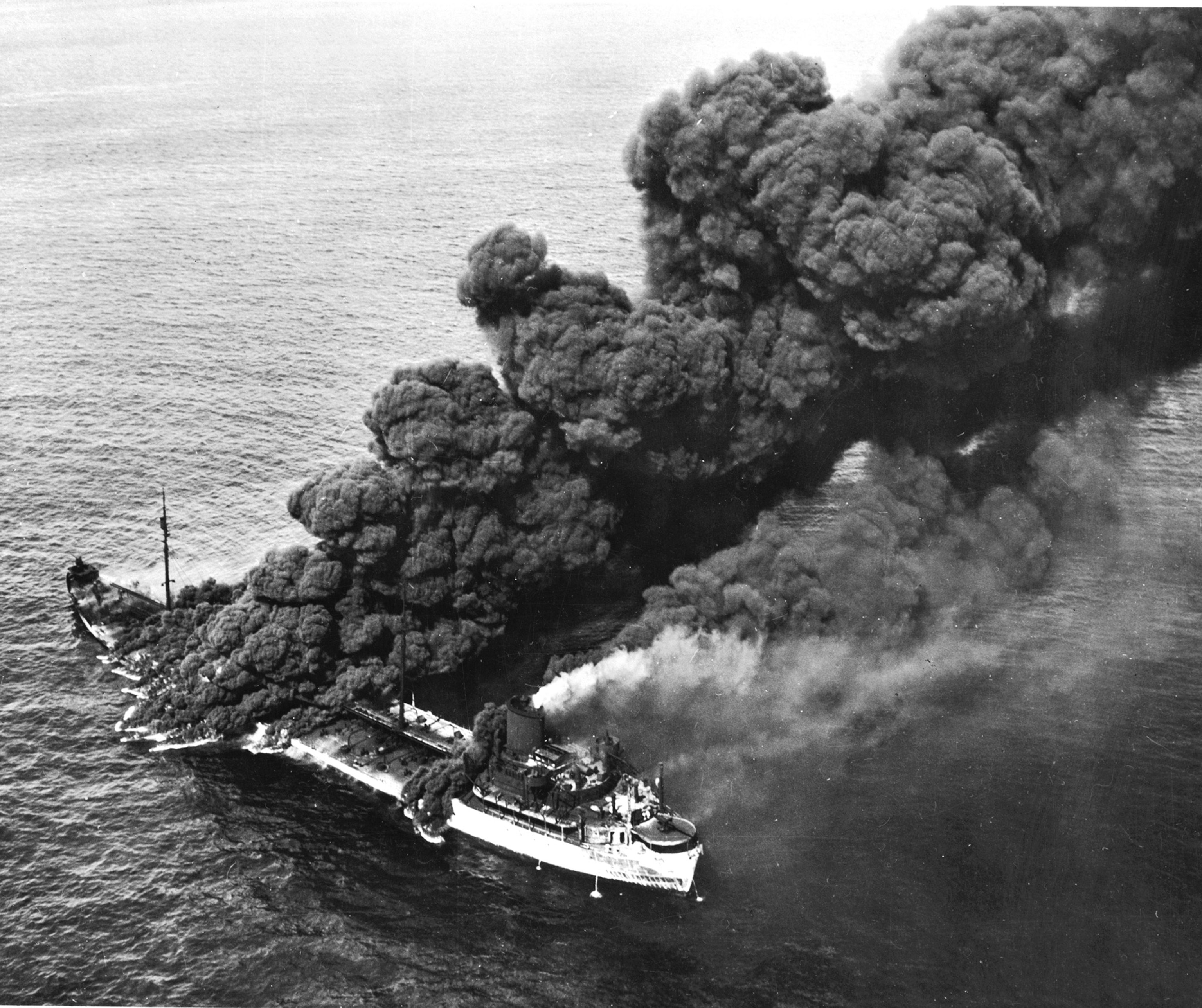 Black And white image dark smoke rises from ship at sea.