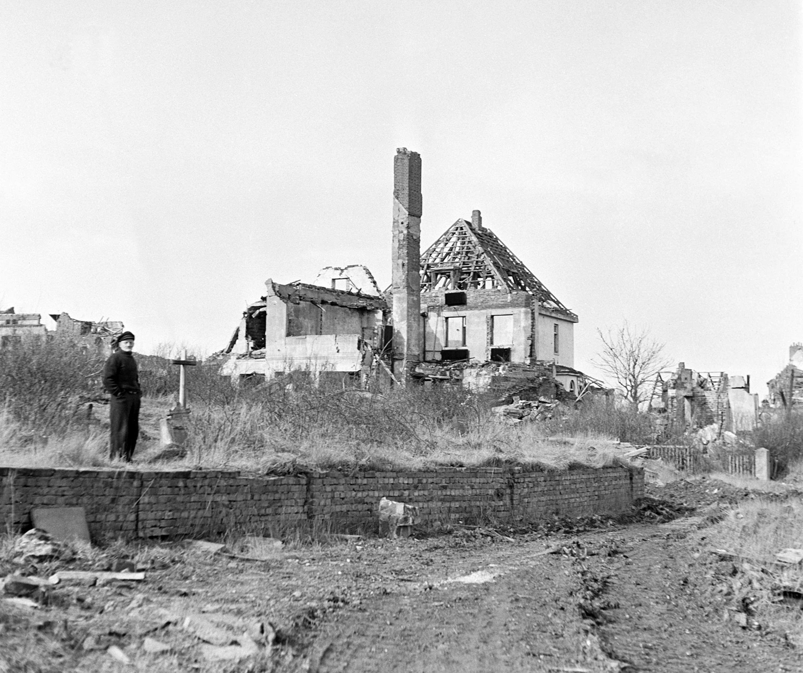 A black and white photo shows a man, standing in front and to the side of a house that has been blown up. Only the frame of the house remains.