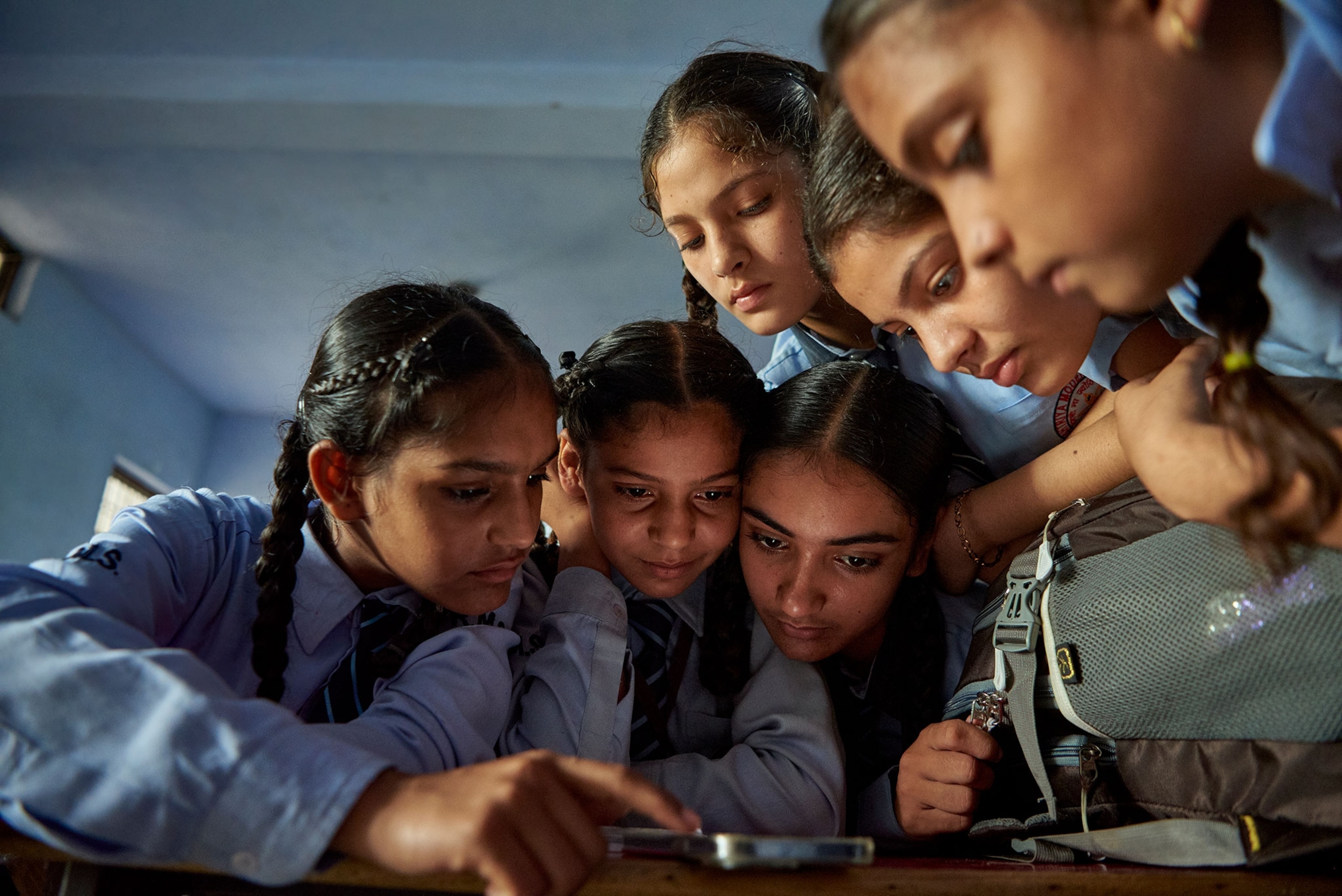 Six young girls dressed in blue school uniforms huddle around a table looking down at a cell phone.