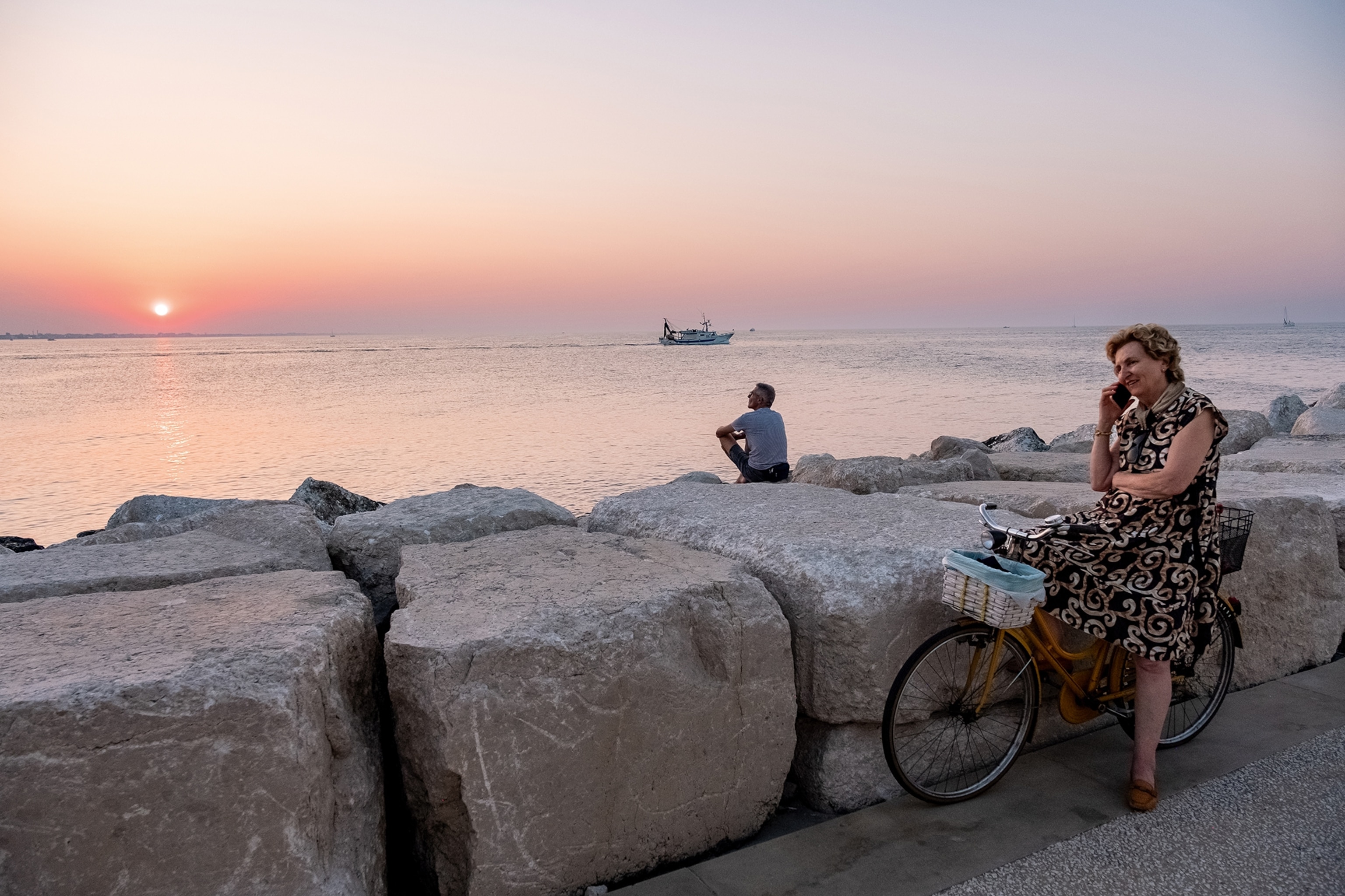 Woman sat on a bike alongside stone walls and the sea at sunset