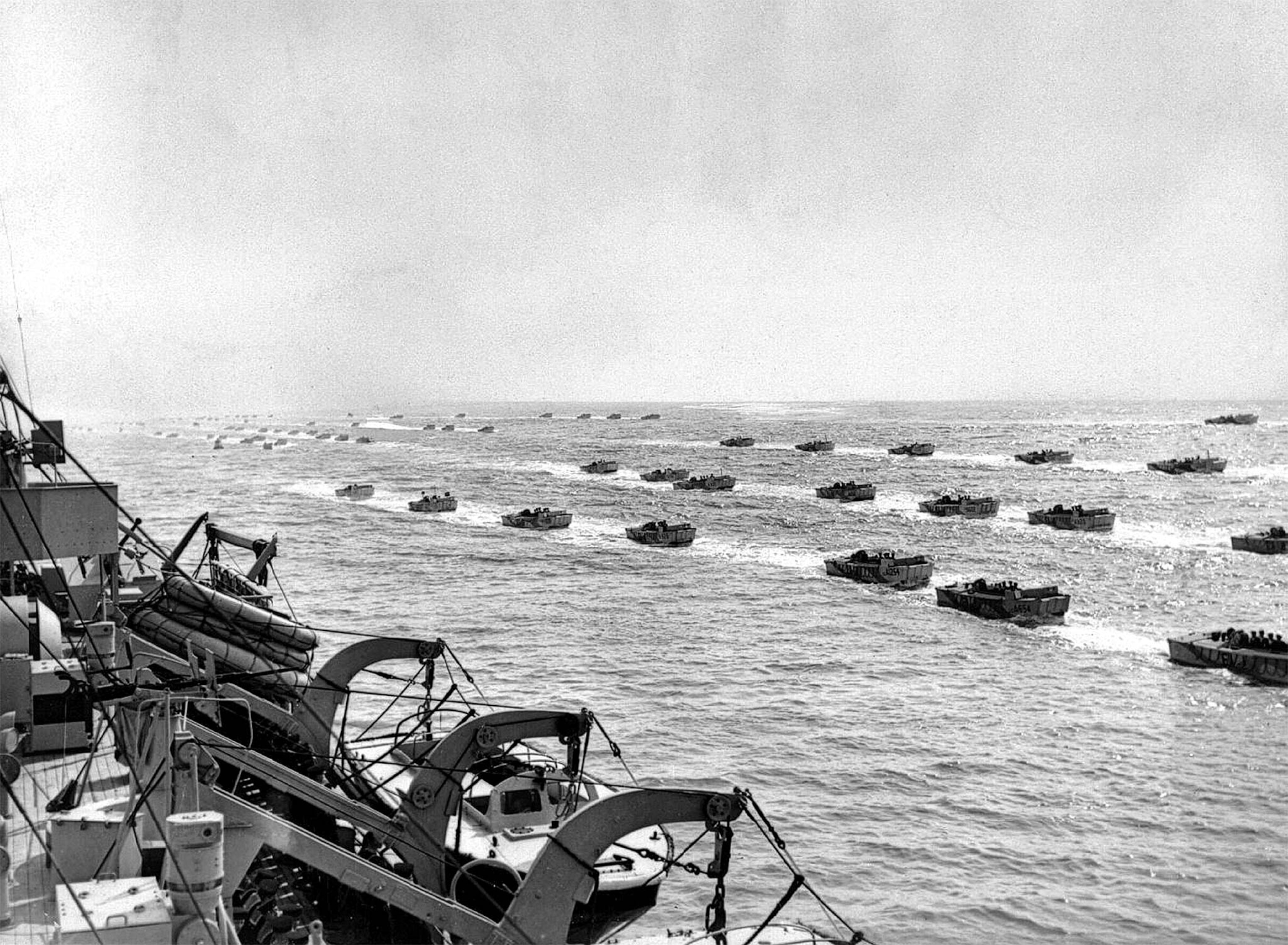 A fleet of landing craft sail in water in a historical photo