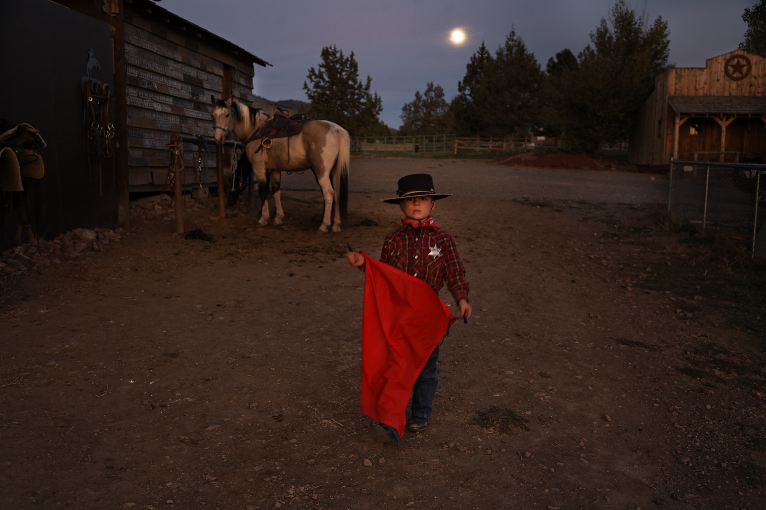 A young boy dressed as a cowboy and holding a red bullfighter's cape standing for a portrait at dusk. A horse looks on from the background and a full moon hangs low on the horizon.