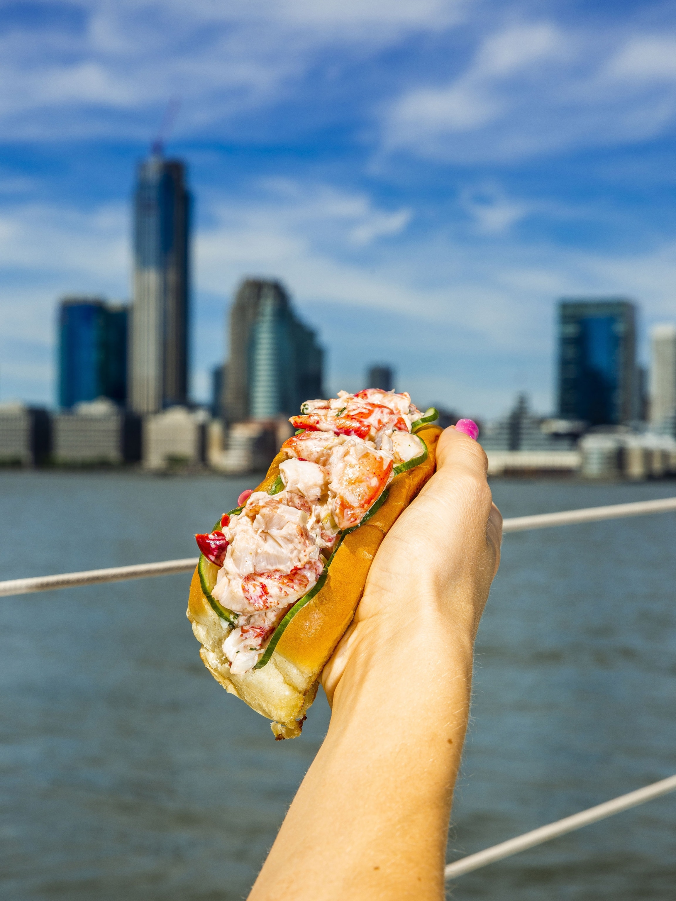 With the Hudson River and skyscrapers of the New York City skyline in the background, a person's outstretched hand holds a lobster roll, complete with golden bun, heaps of pink and red lobster meat, and cucumbers.