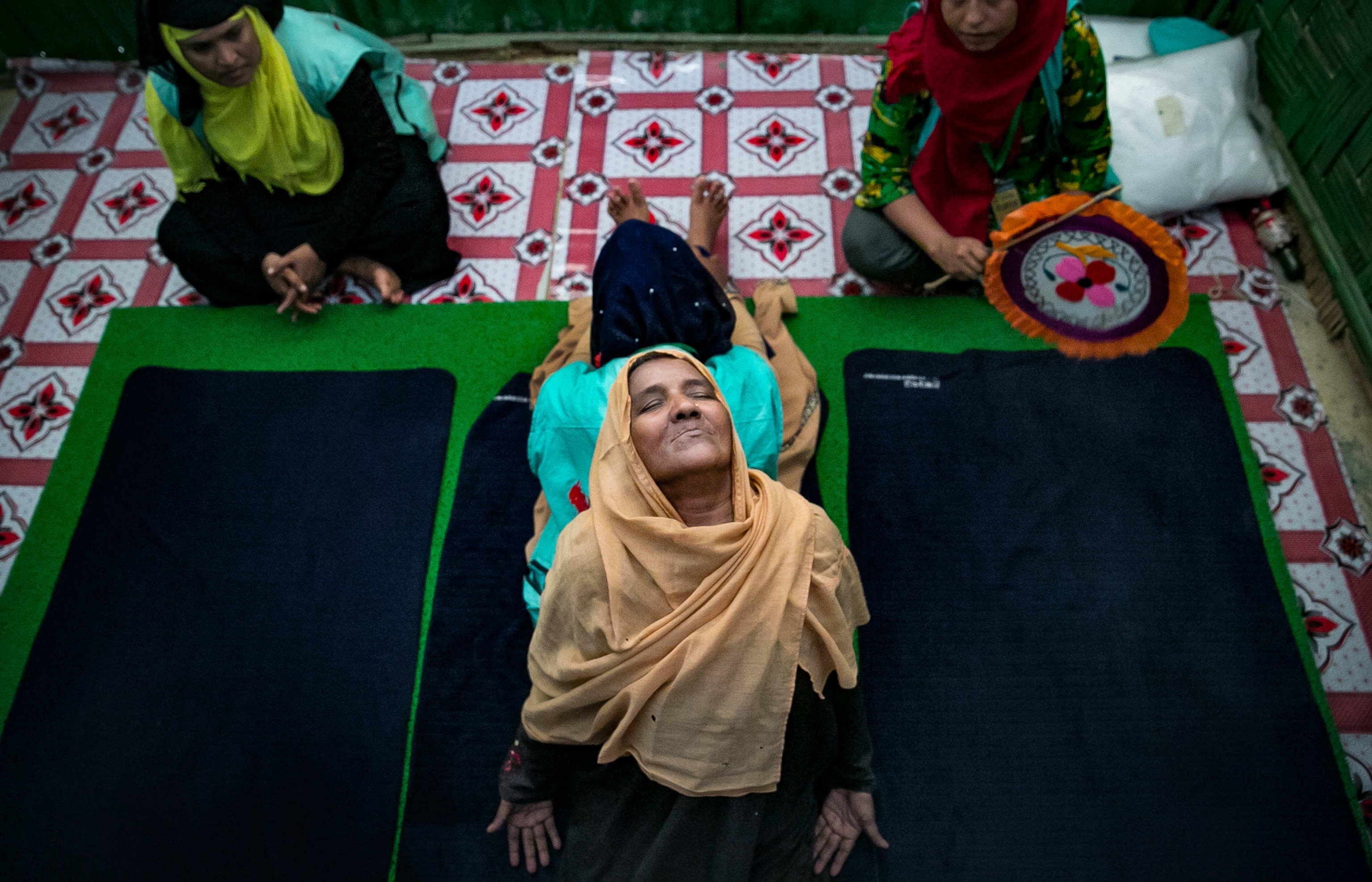 A group of Rohingya women seated on the floor. Two women look on while the other two women are seated back to back as they stretch.