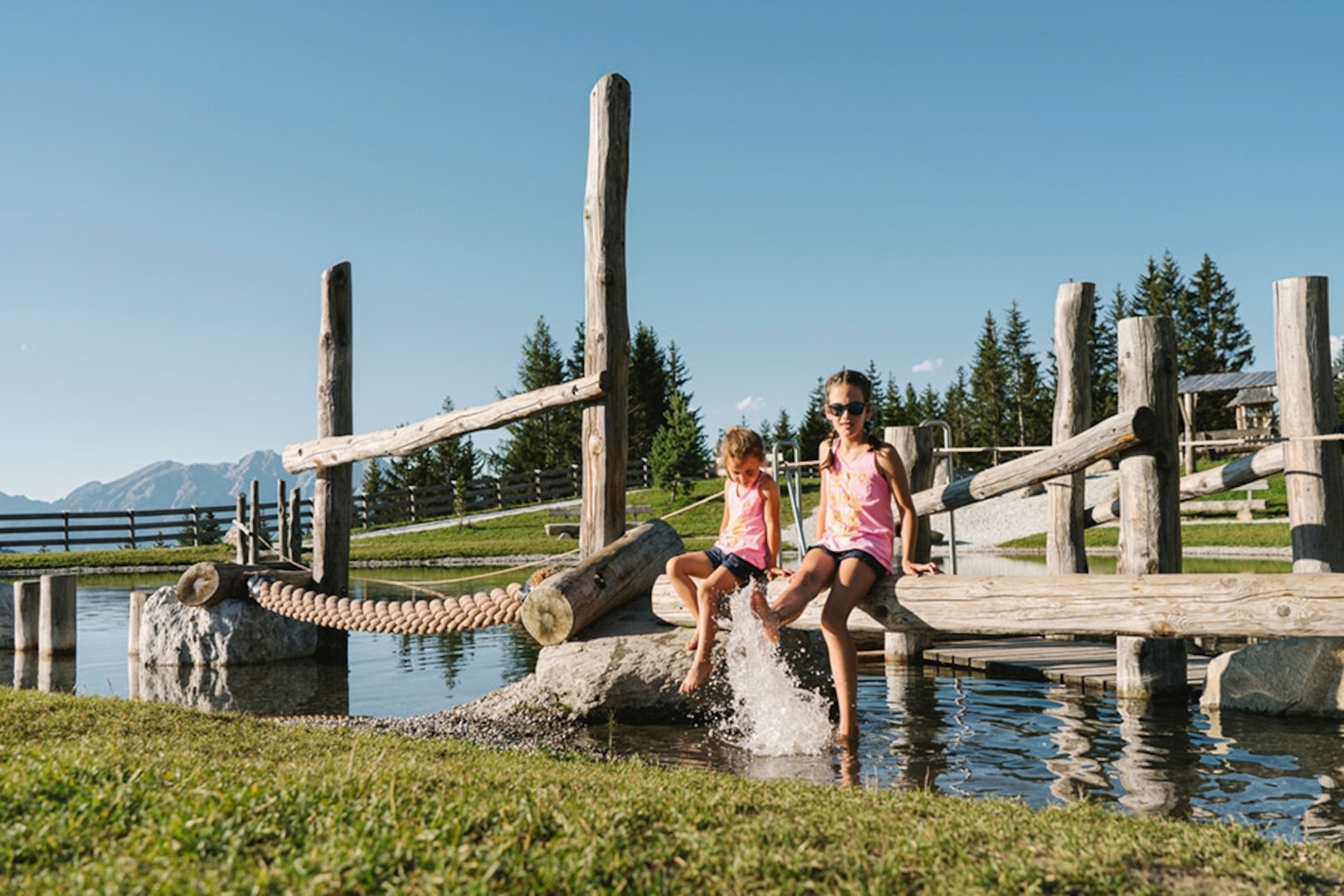 Children sit on a wooden structure kicking their feet in the water