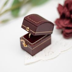 a small wooden box with a ring in it next to a red flower on a lace doily