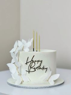 a birthday cake with white flowers and gold candles sitting on top of a table in front of a wall