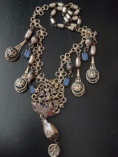 An assemblage featuring a French unmarked antique buckle half with traces of old enamel. It suspends a large baroque freshwater pearl and an antique Victorian cut steel button. The baroque pearl is missing a patch of nacre on the back, but is still luminous and good quality. Accompanying pendants are vintage style blue enameled brass buttons set on brass filigree with faceted iolite, blue Czech glass rectangles and brass capped Czech glass pearls. Finishes with Czech glass pearl chain and hook. Ornate Antique Silver Jewelry With Vintage Charm, Vintage Baroque Jewelry For Formal Occasions, Antique Silver Baroque Jewelry, Antique Silver Baroque Style Jewelry, Antique Gold Decorative Jewelry, Vintage Baroque Gold Jewelry, Ornate Handmade Jewelry For Vintage Occasions, Handmade Ornate Vintage Jewelry, Victorian Vintage Charm Metal Jewelry