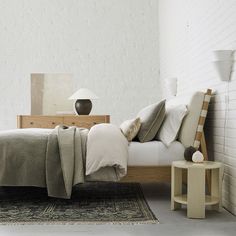 a bed with pillows and blankets on top of it next to a night stand in front of a white brick wall