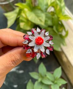 a person holding a red and white brooch in their hand with green plants behind it