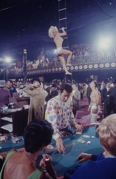 people playing cards at a casino table in front of an audience with one woman on stilts above her head