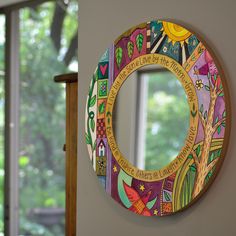 a colorful circular mirror hanging on the wall in front of a window with trees and houses painted on it