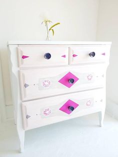 a white dresser with pink and grey designs on the drawers, next to a flower