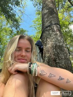 a woman with long hair and tattoos sitting next to a tree in the woods looking at the camera