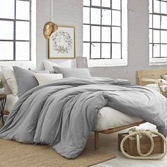 a bed with grey sheets and pillows in a room next to two large windows on the wall