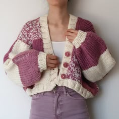 a woman wearing a purple and white cardigan standing next to a wall with her hands on her hips