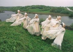 several women in white dresses are walking on the grass near water and trees, with one woman holding her hand up to her head