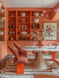 an orange and pink dining room with chairs, table and shelves filled with vases