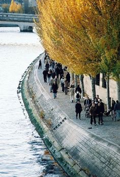 many people are walking on the sidewalk by the water and trees with yellow leaves in autumn