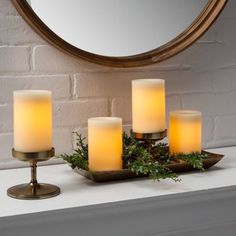 four lit candles sit on a tray with greenery in front of a round mirror