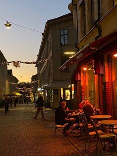 people are sitting at tables on the sidewalk in front of buildings and lights strung across the street