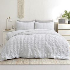 a bed with white comforter and pillows in a room next to a plant on a dresser