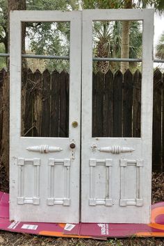 two white double doors sitting next to each other in front of a wooden fence and tree